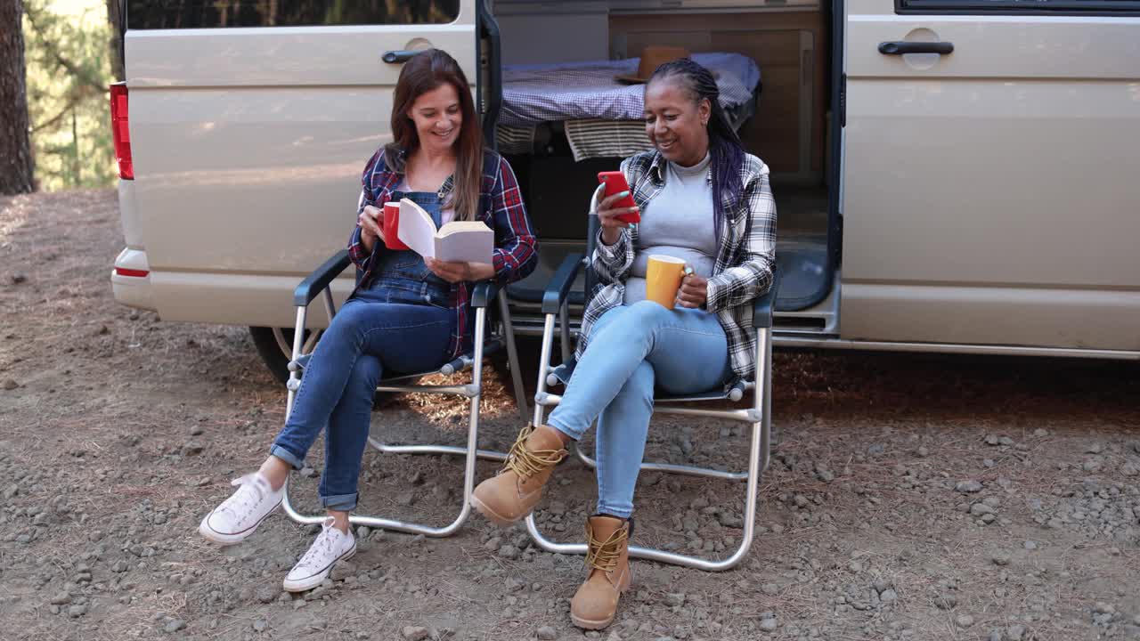 多种族的成熟女性朋友在户外阅读和喝咖啡的同时享受露营车的乐趣-旅行和自然的概念