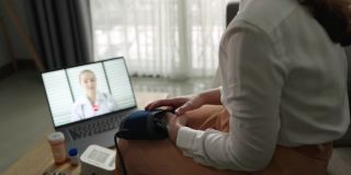 一位亚洲妇女病人在家里用笔记本电脑和她的医生聊天，同时使用血压检查
