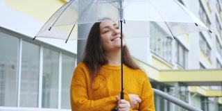 幸福的年轻女子在雨中站在透明的伞下，背景是一幢公寓