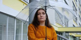 悲伤的女人在雨中站在透明的伞下，背景是一幢公寓