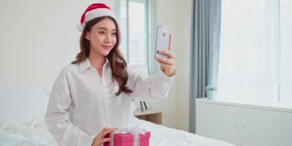 亚洲美女视频通话在线与家人在圣诞节。漂亮的女孩穿着睡衣坐在床上，开心地享受着圣诞礼物，在卧室里和亲戚聊天