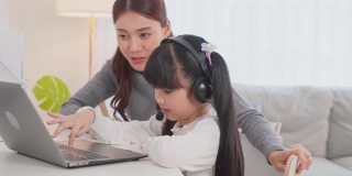 亚洲小女孩和妈妈在家里学习在线课程。学前班的孩子用笔记本电脑做家庭作业，由学校老师通过数字远程互联网在妈妈的支持下完成。