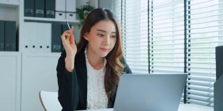 亚洲美丽的商业女性微笑感到快乐，在办公室工作。漂亮的职业女职员穿着正装坐在工作场所的桌子上，在公司使用笔记本电脑享受。