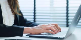 在写字台上用笔记本电脑键盘打字的女商人的双手紧闭。漂亮的职业女职员职员穿着正式的服装坐在工作场所的桌子上，用电脑在办公室工作。