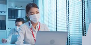 一群亚洲年轻商人在办公室的电脑前工作到很晚。男人和女人戴上防护口罩，保持距离，以新的正常生活理念预防工作场所感染新冠病毒。