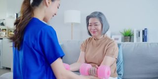 亚洲残疾老妇人做物理治疗师与护士的支持。老年残疾女性坐在客厅的沙发上，用哑铃锻炼身体。