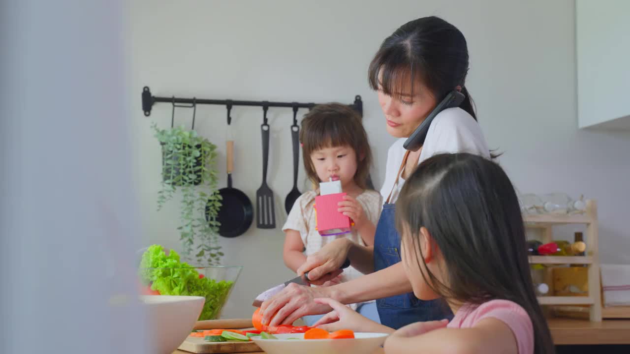 亚洲忙碌的母亲打电话，和女儿在厨房做饭。幸福的家庭，爱美丽的父母，妈妈和小妹妹感到幸福，享受在家里的时间。健康食品的概念