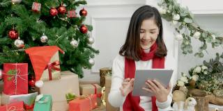 亚洲女人用智能平板电脑坐在圣诞树下的圣诞礼物礼品盒在舒适的客厅。女人手触屏微笑网购，幸福时光圣诞佳节冬季喜庆