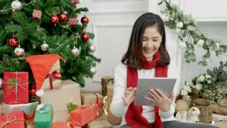 亚洲女人用智能平板电脑坐在圣诞树下的圣诞礼物礼品盒在舒适的客厅。女人手触屏微笑网购，幸福时光圣诞佳节冬季喜庆视频素材模板下载