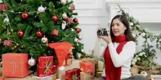 女人摄影师拍摄圣诞装饰球圣诞树在节日冬季庆祝。亚洲妇女在舒适的客厅用相机拍照圣诞树圣诞礼物礼盒