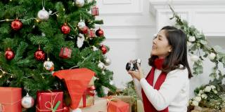 亚洲妇女在舒适的客厅用相机拍照圣诞树圣诞礼物礼盒。女人摄影师拍摄圣诞装饰球圣诞树在节日冬季庆祝