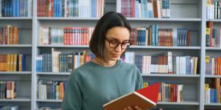 一名年轻的千禧一代女学生站在公共图书馆书架的背景下，在她的笔记本上写作业。一名年轻迷人的女学生的特写照片，背景是大学里的书架