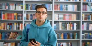 一名年轻的千禧一代学生戴着眼镜，站在图书馆书架的背景下，用智能手机浏览社交网络信息