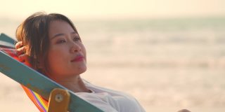 4K美丽的亚洲女人在日落时躺在海边的沙滩椅上