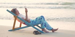 4K美丽的亚洲女人在日落时躺在海边的沙滩椅上