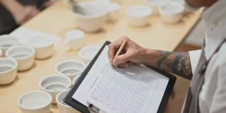 高角度的视角，专业的亚洲华人咖啡师评分者表演咖啡杯，品尝咖啡杯后在剪贴板上写下