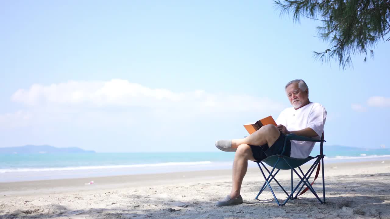 一位亚洲老人坐在海边的户外椅上，享受着看书的乐趣。
