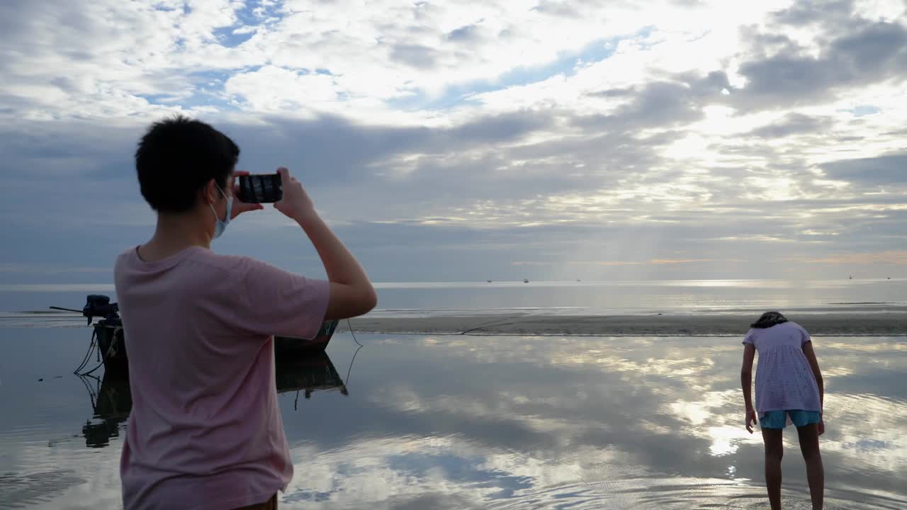 亚洲男孩在早上和家人在海滩上放松时用智能手机拍照，生活理念。