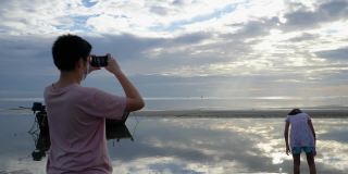亚洲男孩在早上和家人在海滩上放松时用智能手机拍照，生活理念。