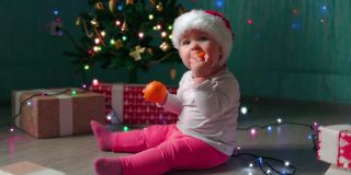 戴着圣诞帽的小女孩坐在圣诞树旁吃着桔子。缓慢的运动。新年假期的概念