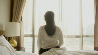 年轻清新的亚洲女性早上穿着睡衣醒来，伸着懒腰从床上起来，手放在大窗户的阳光下，covid - 19封锁结束后的度假旅行视频素材模板下载