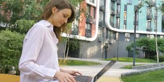 一名年轻女子正在用她的笔记本电脑进行在线会议，一名女商人带着数字平板电脑坐在新现代住宅小区附近的长凳上，在户外远程工作