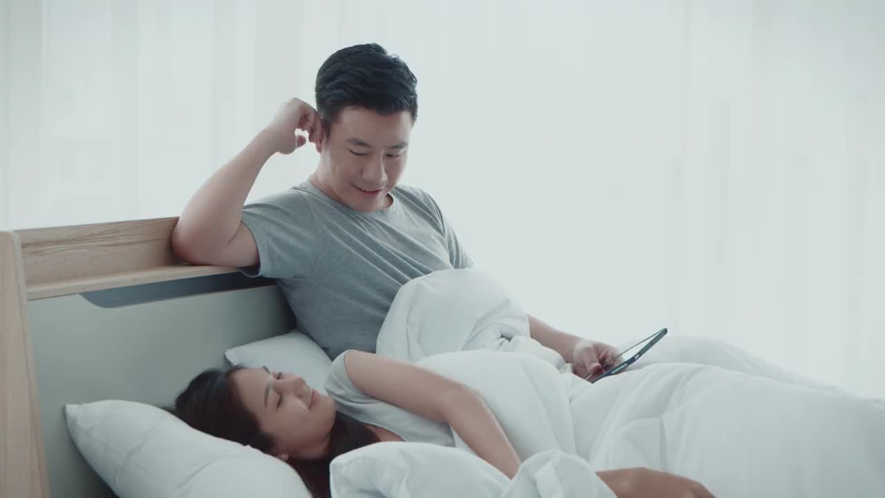 亚洲可爱的已婚年轻夫妇醒来在他们的卧室，使用平板电脑看电影，在睡觉前幸福在一起，4K分辨率，人类关系概念。