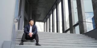年轻的亚洲人沮丧地坐在楼梯上的街道附近的办公商业中心大楼