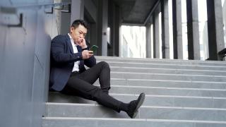年轻的亚洲人沮丧地坐在楼梯上的街道附近的办公商业中心大楼视频素材模板下载