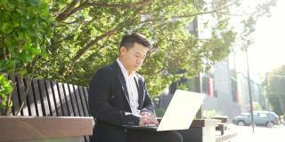 年轻的亚洲商人或自由职业者坐在长椅和工作笔记本电脑在城市公园在现代城市街道
