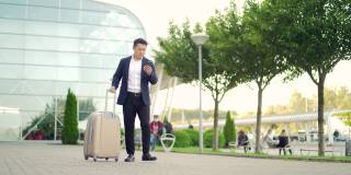 亚洲旅游商人带着行李箱从航站楼走过，背景是一个现代化的城市