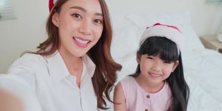 亚洲妈妈带着小孩戴着圣诞老人的帽子和家人在线视频通话。漂亮漂亮的妈妈和年幼的女儿在家里用手机和丈夫聊天庆祝圣诞节