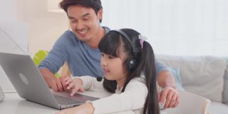 亚洲小女孩和爸爸在家里学习在线课程。学前班的孩子用笔记本电脑做家庭作业，放学后由学校老师通过数字远程互联网在爸爸的支持下完成。
