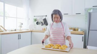 亚洲可爱的家庭，年轻的女儿看着老妈妈在厨房做饭。美丽的女性享受打发闲暇时光，抱着年迈的妈妈在家里的餐桌上烤牛角面包。活动的关系视频素材模板下载