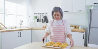 亚洲可爱的家庭，年轻的女儿看着老妈妈在厨房做饭。美丽的女性享受打发闲暇时光，抱着年迈的妈妈在家里的餐桌上烤牛角面包。活动的关系