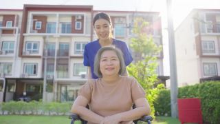 亚洲快乐的老病人奶奶在绿色公园户外玩。护理员微笑着推着坐在轮椅上举起手臂的老人，享受着养老院的炉边护理活动。视频素材模板下载