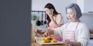 亚洲幸福家庭，年长的妈妈和女儿在家做饭和销售。漂亮漂亮的姑娘用电话接订单，年老年老的母亲把面包装进外卖袋放在桌上。