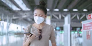 年轻迷人的亚洲女性旅行者戴着口罩，手拿智能手机，查看地图和航班时刻表，在机场航站楼拿着行李和手提包醒来，新的旅行生活方式