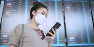 年轻迷人的亚洲女性旅行者戴着口罩，手拿智能手机，查看地图和航班时刻表，在机场航站楼拿着行李和手提包醒来，新的旅行生活方式