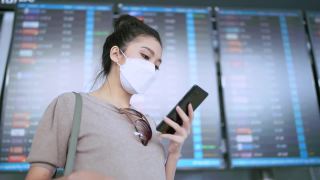 年轻迷人的亚洲女性旅行者戴着口罩，手拿智能手机，查看地图和航班时刻表，在机场航站楼拿着行李和手提包醒来，新的旅行生活方式视频素材模板下载