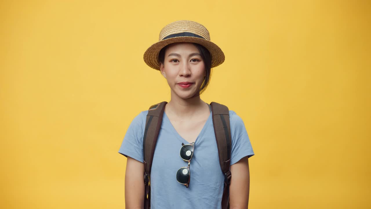 快乐青年亚洲妇女旅行者戴太阳镜帽子和背包微笑看着相机在空白的黄色背景。