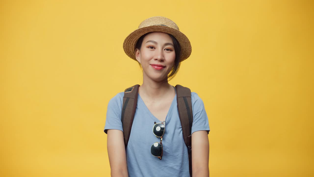 快乐青春美丽亚洲女人旅行者戴帽子和背包显示迷你心与双手爱的标志微笑看着相机在黄色背景。