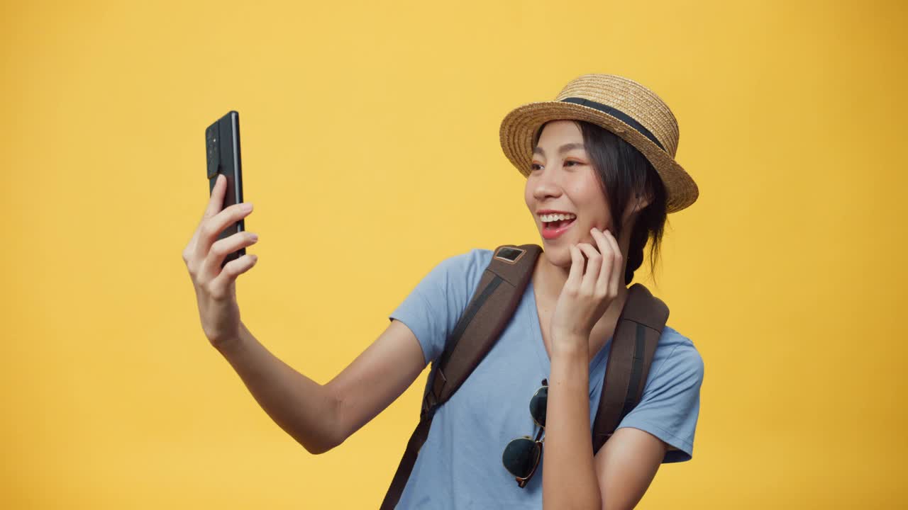 快乐青春美丽亚洲女人旅行者戴太阳镜帽子和背包微笑使用智能手机看相机拍自拍照在空白的黄色背景。