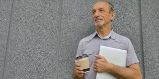 男人的肖像年长的同事与银色笔记本电脑和一杯咖啡等待会议在深灰色的墙壁在早上，远程工作，咖啡休息