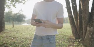 靠近一名身穿白衬衫的男性身体部位，在公园的树荫下散步，放松休闲活动，远程通讯设备，使用无线技术的手