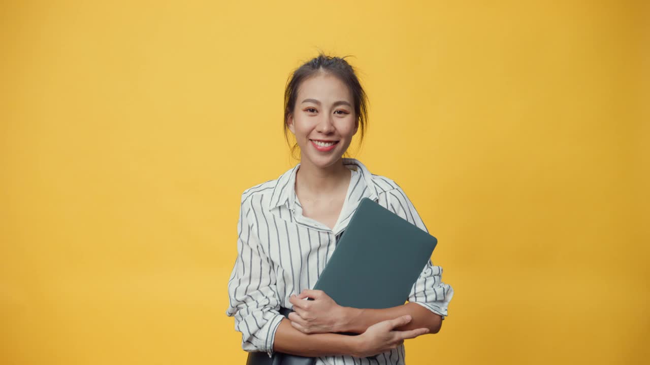 快乐美丽亚洲青春随意的女孩拿着笔记本电脑，站在隔离的黄色空间背景上微笑着看着相机，给出手势呈现广告微笑。