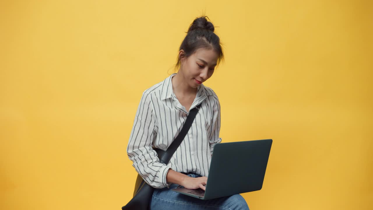 快乐的美女亚洲青春随便的女孩打着笔记本电脑，站在孤立的黄色空间背景上微笑着看着相机，给出手势呈现广告微笑。