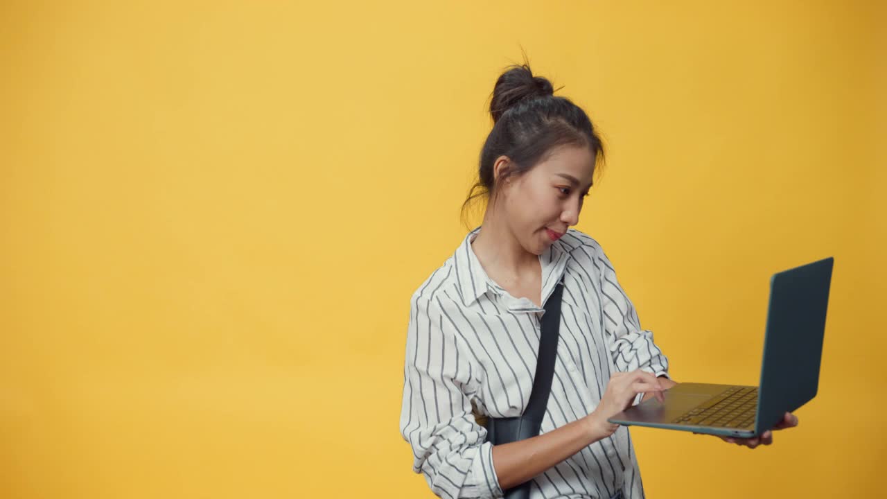 快乐美女亚洲青春休闲女孩打着笔记本电脑，微笑着站在隔离的黄色空间背景下给出标志，展示成功购买网上广告。