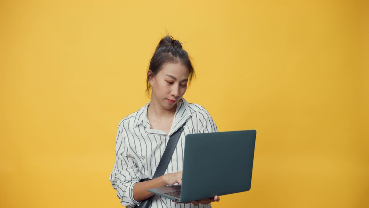 快乐的美女亚洲青春随便的女孩打着笔记本电脑，站在孤立的黄色空间背景上微笑着看着相机，给出手势呈现广告微笑。