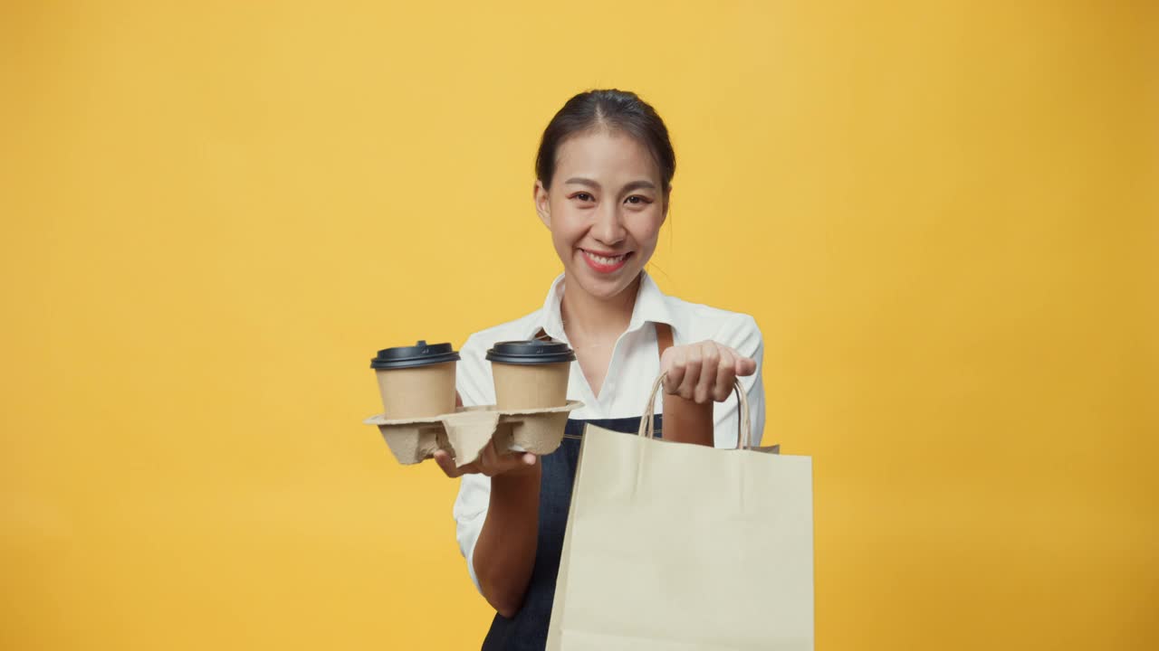 快乐的微笑亚洲女服务员穿着围裙友好地看着镜头给顾客咖啡或茶和美味的食品在手工纸袋隔离在黄色背景。